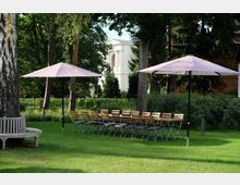 Eine grüne Parklandschaft mit Tischen und Stühlen, die unter zwei großen, lilafarbenen Sonnenschirmen arrangiert sind. Im Hintergrund sind hohe Bäume und ein helles, klassizistisches Gebäude mit Säulen und Rundbogenfenstern zu sehen.