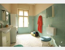 Badezimmer mit türkisfarbenen Wandfliesen und weißen Bodenfliesen. Im Raum befinden sich eine Toilette, ein Waschbecken, eine Waschmaschine, ein Fenster mit Holzrahmen sowie ein an der Wand aufgehängter roter Bademantel.