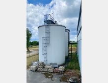 Ein großer, zylindrischer Industriesilo mit weißer Außenverkleidung und einer befestigten Metallleiter an der Seite. Im Vordergrund stapeln sich Pflastersteine auf Paletten, und im Hintergrund sind Grünflächen und ein Zaun zu sehen.