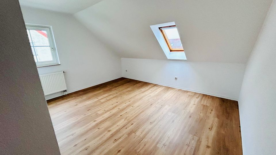 Leerer Dachgeschossraum mit schrägen weißen Wänden und hellem Holzfußboden. Ein normales Fenster mit Heizkörper darunter sowie ein Dachfenster sorgen für Tageslicht.