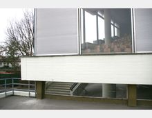 Ansicht eines modernen Gebäudes mit breiten Fensterfronten. Im Inneren sind Reihen von hellbraunen Sitzplätzen sichtbar, darunter befindet sich ein überdachter Eingang mit einer Treppe, umgeben von Betonstützen.