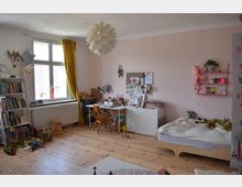 Helles Kinderzimmer mit Holzfußboden und pastellrosanen Wänden, ausgestattet mit einem Einzelbett, einem Schreibtisch mit Stuhl und Regalen mit Büchern und Spielsachen. Ein großes Fenster mit gelbem Vorhang lässt Tageslicht herein, und eine dekorative Deckenlampe hängt zentral im Raum.