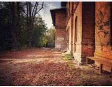 Backsteingebäude mit sichtbaren Graffiti und einer Bank an der rechten Seite, davor ein mit Herbstlaub bedeckter gepflasterter Weg, der zu einer baumreichen Umgebung führt. Ein Schild mit der Aufschrift „Fahrkarten-Ausgabe“ ist an der Gebäudewand angebracht.