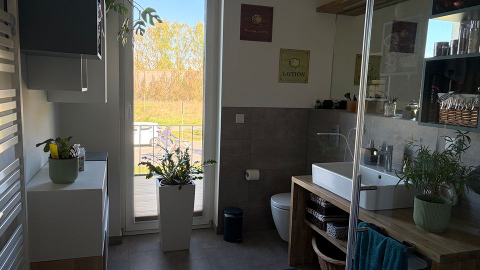 Modernes Badezimmer mit grauen Fliesen, großem Fenster bis zum Boden und Blick ins Freie. Rechts ein Waschtisch aus Holz mit offenem Regal, Wandregal mit Pflegeprodukten und Pflanzen sowie ein WC; vorne ist die Glaswand einer Dusche sichtbar.