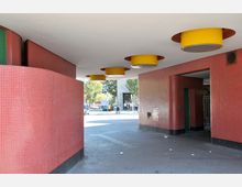 Überdachter Durchgang mit roten Fliesenwänden und gelben, runden Deckenelementen. Im Hintergrund führen der Durchgang und die Straße zu einem offenen, städtischen Bereich mit Bäumen und parkenden Autos.