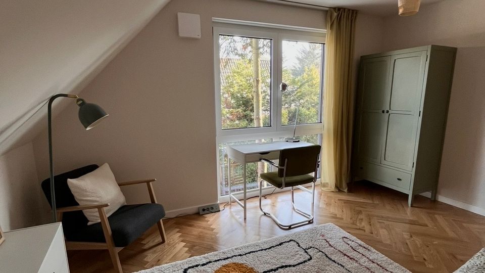 Ein gemütliches Schlafzimmer mit schräger Decke, Holzboden im Fischgrätmuster und einem großen Fenster mit gelbem Vorhang, das auf einen Garten blickt. Der Raum enthält ein Bett, eine Kommode, einen Sessel mit Stehlampe, einen kleinen Schreibtisch mit Stuhl und einen grünen Kleiderschrank.