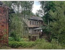 Verfallenes Gebäude inmitten dichter Vegetation, mit einer Fassade aus verwittertem Putz und mehreren zerbrochenen Fenstern; links im Bild ist ein Teil einer roten Backsteinmauer sichtbar. Im Vordergrund befinden sich Büsche und hohes Gras, während Bäume die Umgebung dominieren.