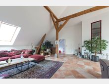 Ein geräumiges Wohnzimmer mit schräger Decke und sichtbaren Holzbalken. Der Raum hat ein großes Dachfenster, eine rote Couch mit passenden Ottomanen, einen bunten Teppich auf Terrakottafliesen, verschiedene Pflanzen und eine offene Tür zu einem angrenzenden Raum.