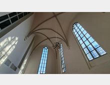 Innenraum einer Kirche mit weiß verputzten Wänden, hohem Kreuzrippengewölbe und großen, schmalen Spitzbogenfenstern mit Bleiverglasung. Von der Decke hängt ein moderner, kugelförmiger Kronleuchter.