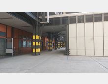 Industriehalle mit einem Mix aus Metall- und Betonstrukturen, schwarz-gelbe Pfeiler markieren den Durchgangsweg. Linke Seite mit orangefarbenen Wänden und Fenstern, rechte Seite mit einer grauen Wand aus Platten.