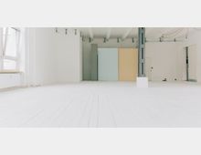 Minimalistischer Innenraum mit weiß gestrichenen Holzdielen und hohen Decken, linkerhand ein großes, teilweise geöffnetes Fenster. An der Rückwand stehen senkrechte Tafeln in Pastellfarben (Blau und Gelb), die durch eine Metallstütze rechts ergänzt werden.