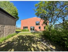 Backsteingebäude mit großen Fenstern in einem Gartenbereich, umgeben von Bäumen und Grünflächen; links im Vordergrund eine Wand aus bräunlichem Putz mit Gras am Boden.