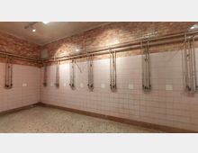 Ein leerer Duschraum mit rosa Fliesenwänden und mosaikartigen Fliesen auf dem Boden. An den Wänden befinden sich freiliegende Wasserrohre und mehrere Duscharmaturen unterhalb von Verkabelungen.