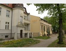 Straßenansicht einer historischen Pension mit hellgelber und cremefarbener Fassade, kleinem Turm, dekorativen Stuckelementen und einem seitlich angebauten Gebäude. Im Vordergrund befindet sich ein gepflasterter Gehweg, umgeben von Rasenflächen und Bäumen.