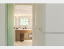 Blick in ein modernes Badezimmer mit einer weißen Wand im Vordergrund. Im Hintergrund befinden sich ein Waschtisch mit Holzverkleidung, ein dekorativer Hocker und ein Fenster mit Sprosseneinteilung, durch das grüne Bäume zu sehen sind.