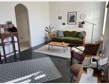 Wohnzimmer mit einer grünen, gepolsterten Vintage-Couch, einem kleinen Holztisch und einem roten Sessel. Im Raum befinden sich ein schlichter Schreibtisch mit Schreibmaschine, ein Bücherregal, mehrere Bilder an der weißen Wand und eine stehende Lampe; der Boden ist teilweise mit Teppichen bedeckt.