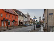 Straßenszene in einer historischen Stadt mit farbigen Altstadthäusern, Bürgersteig und parkenden Autos; im Hintergrund eine Kirche mit barockem Glockenturm. Straßenschilder und Laternen befinden sich entlang der gepflasterten Gehwege.