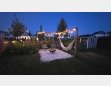Garten-Lounge im Wohngebiet bei Dämmerung mit Holzpergola und Lichterkette über einer Kiesfläche, darauf eine Hängematte, Sitzmöbel und eine niedrige Feuerstelle. Umgeben von Rasen, Blumenbeeten und Sträuchern, rechts mit kleinem Gartenhaus.