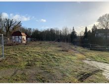 Grasbewachsene Freifläche mit spärlicher Vegetation, im Hintergrund ein kleines Fachwerkhäuschen mit rotem Dach sowie einige Bäume und Sträucher; rechts sind Holzkonstruktionen und ein Gebäude mit Fachwerkelementen sichtbar. Der Himmel ist hell mit vereinzelten Wolken.