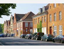 Straßenszene mit einer Reihe von historischen Backsteinhäusern mit roten Dächern und Sprossenfenstern. Am Straßenrand sind geparkte Autos, während ein Radfahrer auf der Fahrbahn unterwegs ist, und im Hintergrund ist leichtes Grün von Bäumen zu erkennen.