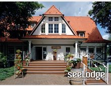 Außenansicht eines Gebäudes mit weißer Fassade, roten Dachziegeln und einem Giebeldach, das als Restaurant und Hotel ("seeLodge") dient. Der Eingangsbereich ist durch eine Holzveranda mit Treppe und Blumendekorationen eingerahmt.