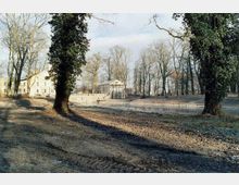 Parklandschaft mit großen, teils mit Efeu bewachsenen Bäumen, einem kleinen Gebäudepavillon im klassizistischen Stil am Rand eines Teiches und einem größeren Gebäude im Hintergrund. Der Boden ist teils unbefestigt, mit Reif bedeckt, und die Umgebung wirkt herbstlich oder winterlich.