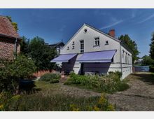 Weißes Gebäude mit seitlichem Satteldach, zwei großen, lila Markisen über einer Terrasse mit Gartenmöbeln. Umgeben von üppigem Grün, Kiesweg und einem angrenzenden roten Ziegelhaus.