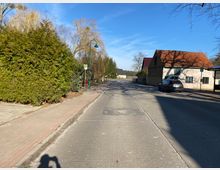 Straßenansicht in einem ländlichen Gebiet mit einem Gehweg, einer Bushaltestelle und Laternen auf der linken Seite. Rechts sind ein Haus mit rotem Ziegeldach und ein parkendes Auto zu sehen, umgeben von Bäumen und Sträuchern unter klarem Himmel.