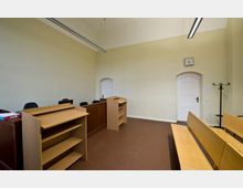 Ein schlichter Gerichtssaal mit hellbeigen Wänden, Holzmöbeln, darunter ein Richterpult, Zuschauerbänke und ein Rednerpodium. Ein weißes, geschwungenes Türportal und eine analoge Wanduhr befinden sich an der Rückwand.