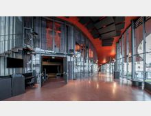 Moderner Veranstaltungsraum mit gebogener Glasfassade und einem roten Boden sowie einer roten Decke. Er verfügt über große Fenster, die den Blick auf die Umgebung ermöglichen, und eine offene Bühne im Hintergrund.