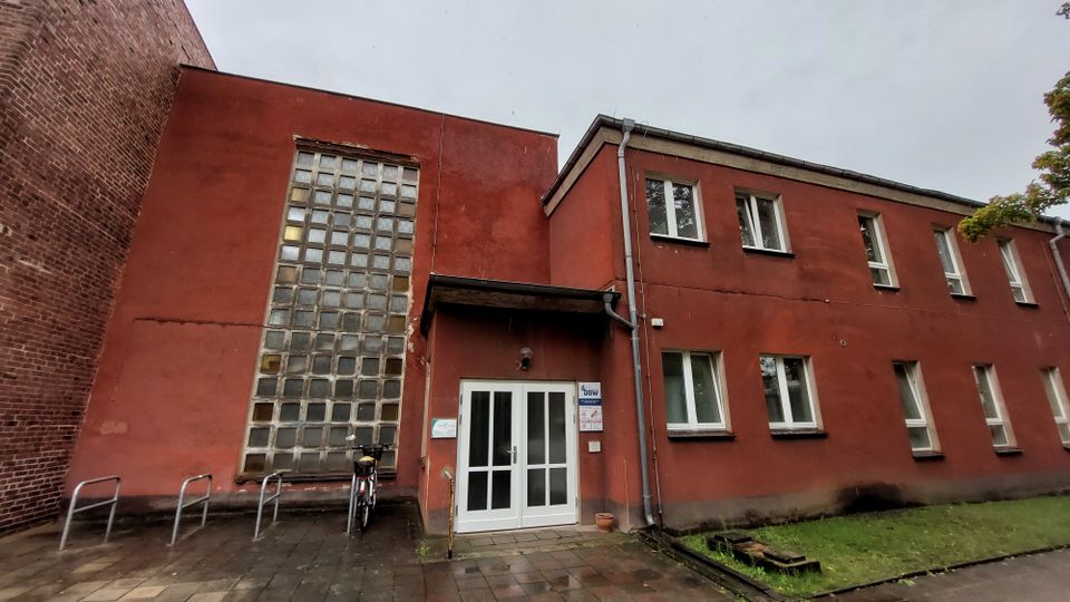 Zweistöckiges Gebäude mit roter Fassade und Flachdach, großen Glasbausteinfenstern auf der linken Seite und mehreren rechteckigen Fenstern auf der rechten Seite. Vor dem Eingang mit weißen Doppeltüren befinden sich Fahrradständer und ein gepflasterter Weg, daneben ein kleiner Grünbereich.