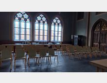 Ein Raum mit hohen, farbigen gotischen Fenstern, Reihen von Stühlen in einem Konferenz-Setup und einem Tisch im Hintergrund, gedeckt mit Gläsern, Tassen und Getränken. Rechts im Bild sind verzierte Holztüren und ein dekorativer Schrank sichtbar.