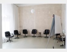 Ein heller, minimalistisch eingerichteter Besprechungsraum mit einer Reihe schwarzer Drehstühle, einem kleinen Hocker und einem Flipchart. Die Wände sind schlicht und hell gehalten, mit großen weißen Vorhängen an der Fensterseite.