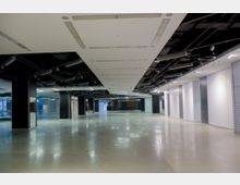 Großer, leerer Innenraum mit poliertem Boden, schwarzen Säulen und einer weißen abgehängten Decke mit sichtbaren schwarzen Lüftungselementen. Der Raum hat helle Wände und ist von Deckenleuchten gleichmäßig ausgeleuchtet.