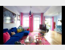 Wohnzimmer mit moderner und bunter Einrichtung. Ein großes blaues Sofa mit bunten Kissen, ein gemusterter Teppich und eine Glas-Couchtischgruppe bestimmen den Raum; im Hintergrund sorgen rosa Vorhänge, hohe Fenster und eine offene Küchenzeile mit blauer Theke für Helligkeit und Farbige Harmonie.