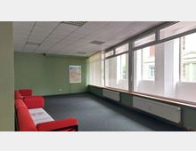 Langer Warteraum mit grünen Wänden, einer Fensterfront mit vertikalen Jalousien und einem Heizkörper darunter. Links stehen rote Sofas, und an der Wand hängt eine bunte Karte.