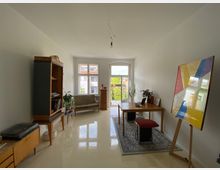 Heller Wohnraum mit minimalistischem Einrichtungsstil, unter anderem mit einem Holztisch, einem Sofa, Pflanzen und einem modernen Kunstwerk auf einer Staffelei. Große Fenster und eine Balkontür im Hintergrund lassen viel Tageslicht herein und bieten Ausblick auf benachbarte Gebäude.