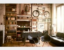 Industriell eingerichteter Raum mit Holzboden, einer großen Bücherwand, einem an der Wand befestigten Fahrrad und Dekorationen wie alten Flaschen und Retro-Gegenständen. Im Vordergrund stehen ein schwarzer Kickertisch, zwei schwarze Stühle und ein Ledersofa, während ein großes Fenster Tageslicht hereinlässt.