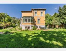 Mehrstöckige gelbe Backsteinvilla mit großen Fenstern und verglastem Erker, umgeben von einem weitläufigen Garten mit Rasenfläche, Sträuchern und hohen Bäumen. Das Gebäude hat einen hell abgesetzten Sockel und steht vor blauem Himmel in einer grünen, parkähnlichen Umgebung.