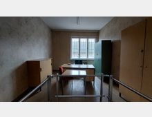 Ein einfacher Büroraum mit einem Schreibtisch und zwei Stühlen, umgeben von Holzschränken und einem grünen Metallschrank. Das Zimmer hat ein Fenster mit Gitterstäben, durch das das Dach eines benachbarten Gebäudes sichtbar ist, sowie eine helle Tapete mit einem dezenten Muster.