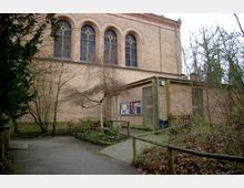 Außenansicht eines Backsteingebäudes mit großen, dekorativen Rundbogenfenstern. Im Vordergrund befindet sich ein kleiner Weg, der mit Geländern umgeben ist, sowie ein bepflanztes Beet und kahles Geäst von Bäumen und Sträuchern.