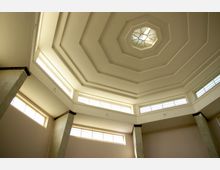 Innenraum mit achteckiger Decke und zentralem Oberlicht mit geometrischem Muster, umgeben von mehreren rechteckigen Fenstern, die Tageslicht hereinlassen. Betonpfeiler an den Wänden stützen die Struktur.