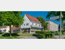 Straßenecke mit einem hellen, zweistöckigen Wohn- und Geschäftshaus mit rotem Satteldach; im Erdgeschoss befinden sich Läden und ein Café mit Außensitzplätzen. Im Hintergrund weitere Wohngebäude, umgeben von Gehwegen, Grünflächen und Bäumen. Ein Straßenschild mit der Aufschrift „Andrézeile“ ist im Vordergrund sichtbar.