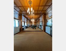 Langer, eleganter Flur mit holzvertäfelter Decke und Wänden, dekorativen Kronleuchtern und Teppichboden. Links und rechts befinden sich verglaste Büroräume mit matten Sichtschutzstreifen, und in der Mitte stehen mehrere Pflanzen.