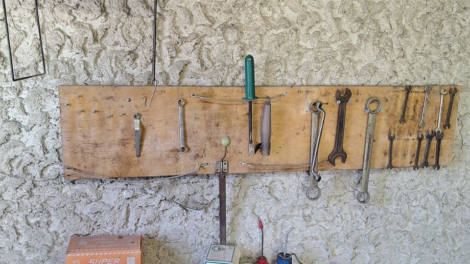 Werkstattwand aus rau verputztem Stein mit zwei Holzleisten als Werkzeughalter, an denen mehrere Schraubenschlüssel und Handwerkzeuge hängen. Davor eine staubige Werkbank mit verschiedenen Behältern, einer Kabelrolle und einer großen Metallsäge bzw. Schneidmaschine.