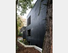 Modernes Gebäude mit einer Fassade aus schwarzem Holzpaneel, gelegen in einer waldartigen Umgebung mit Bäumen und Laub am Boden. Große rechteckige Fenster unterbrechen die glatte Architektur, die von Betonfundamenten umgeben ist.