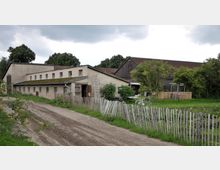 Ein Bauernhof mit mehreren Gebäuden aus verputztem Beton und Holz, flankiert von Bäumen und einem niedrigen Holzzaun. Im Vordergrund verläuft ein unbefestigter Weg, und auf der rechten Seite steht ein Fahrrad sowie ein kleiner Garten mit Möbeln und Pflanzen.