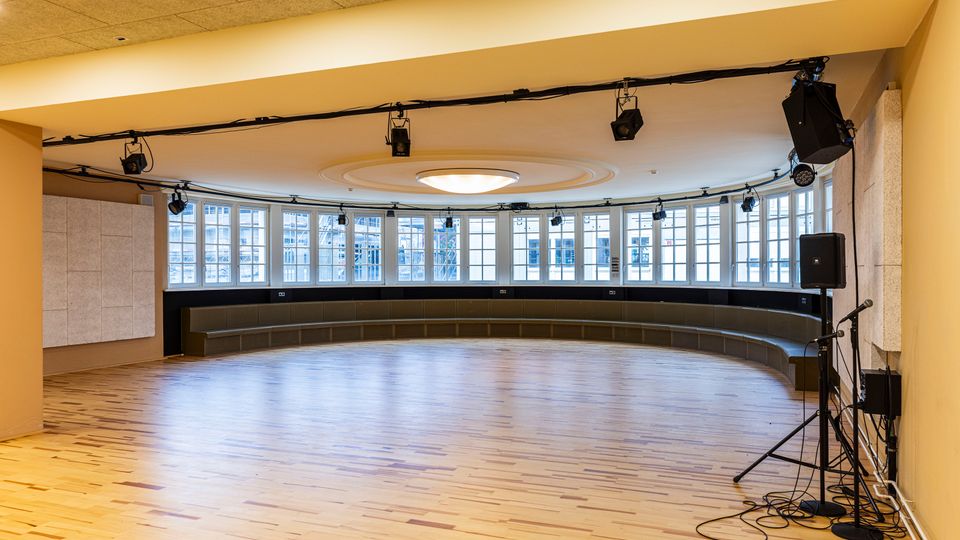 Großer, leerer Veranstaltungs- oder Probenraum mit hellem Holzboden, gebogener Fensterfront und umlaufender Sitzbank entlang der Wand. An der Decke sind Beleuchtungs- und Tontechnik montiert, rechts stehen Lautsprecher und Mikrofonstative.