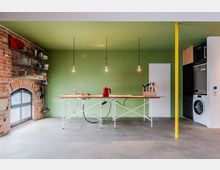 Offene, minimalistische Küche mit grüner Wand, zentraler Kücheninsel aus Holz mit Spüle und drei hängenden Glühbirnenlampen. Links eine freigelegte Ziegelwand mit Bogenfenster und offenen Regalen, rechts ein Geräteschrank mit Waschmaschine sowie eine gelbe Stützsäule auf Betonboden.
