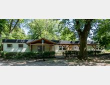 Ein flaches Gebäude mit hellem Anstrich und schwarzem Dach, umgeben von Bäumen und Büschen. Vor dem Gebäude befindet sich ein überdachter Eingangsbereich mit Holzbalken, Sitzgelegenheiten und ein Baum im Vordergrund.